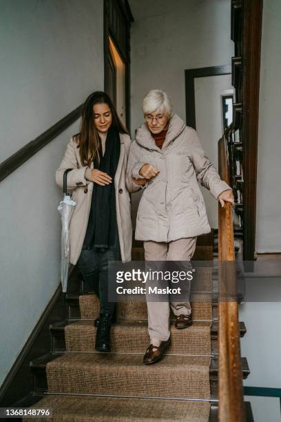 senior woman taking support of female caregiver while moving down on steps at home - holding umbrella stock pictures, royalty-free photos & images