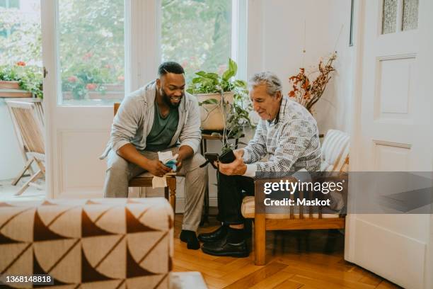 senior man sharing smart phone with male healthcare worker sitting in living room - carer allowance stock pictures, royalty-free photos & images
