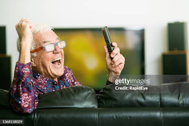 happy elderly man punches the air in joy as he sits watching tv and holding remote control - big toothy smile stock pictures, royalty-free photos & images