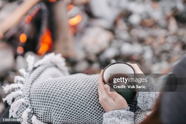 camper holding cup of tea next to campfire - wrapped in a blanket stockfoto's en -beelden