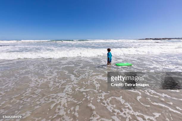 boy (8-9) with surfboard on beach - ankle deep in water stock pictures, royalty-free photos & images