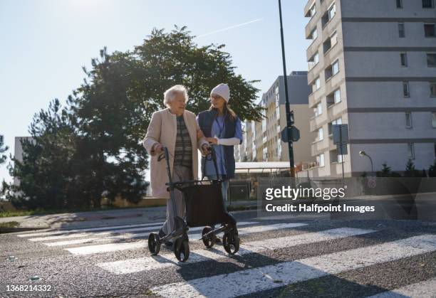 senior woman and caregiver outdoors on a walk with walker in town, crossing the road. - healthcare worker stock-fotos und bilder