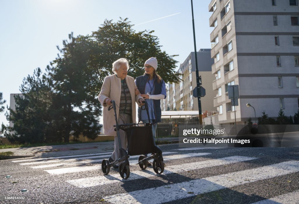Senior woman and caregiver outdoors on a walk with walker in town, crossing the road.
