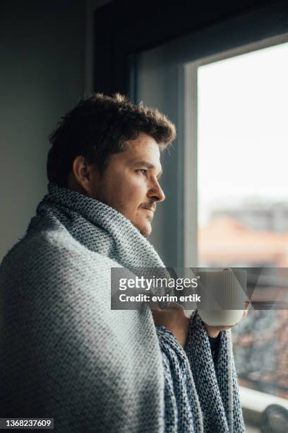 sick man with blanket looking through the window with a cup of tea - winter illness stock pictures, royalty-free photos & images