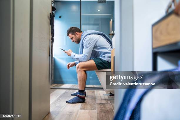 young man with shirt, sitting on the toilet, holding a mobile phone and having online conversation in a difficult situation - pants down bildbanksfoton och bilder