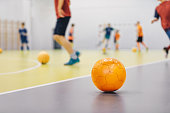 Orange Futsal Soccer Ball on Indoor Training Pitch. Young Players On Sports Practice Running Balls in Blurred Background. School Sports Class For Kids