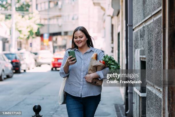 una donna sorridente in sovrappeso che legge un messaggio sul suo cellulare mentre tiene in mano un sacchetto di carta con la spesa nell'altra mano - grasso nutrienti foto e immagini stock