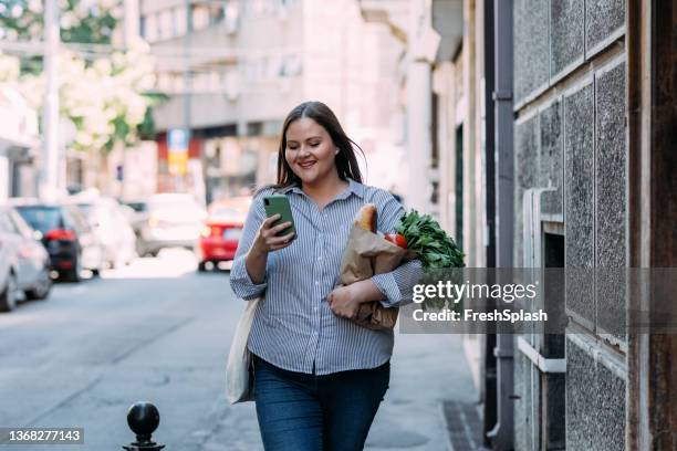 a smiling overweight woman reading a message on her mobile phone while holding a paper bag with groceries in the other hand - overweight bildbanksfoton och bilder