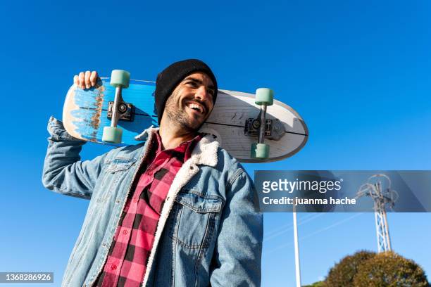man carrying his skateboard on his shoulders while walking with a smile in the park. - mens free skate imagens e fotografias de stock
