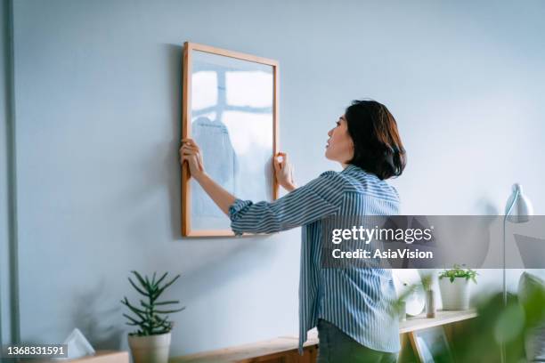 joven asiática decorando y colocando un marco de fotos en la pared de la casa - cuadro colgando fotografías e imágenes de stock