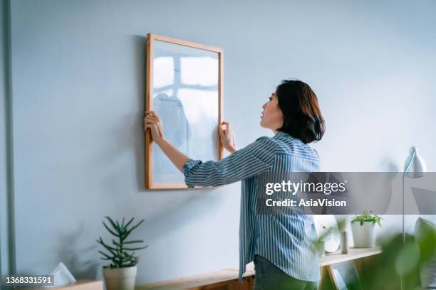 joven asiática decorando y colocando un marco de fotos en la pared de la casa - decorar fotografías e imágenes de stock