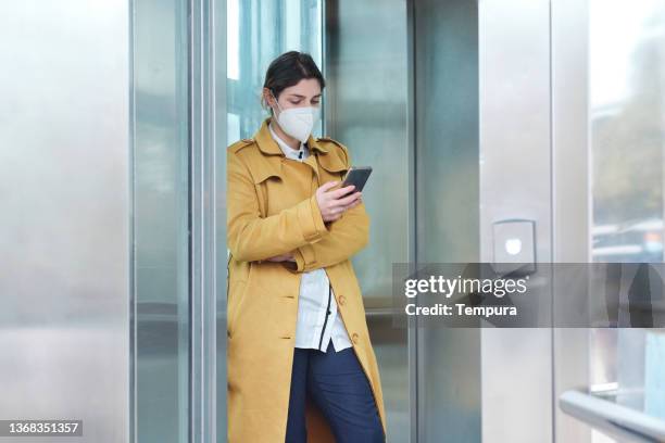 a businesswoman stands in an elevator looking at a mobile phone. - social distancing elevator stock pictures, royalty-free photos & images