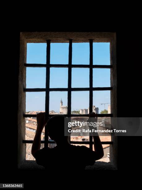 young teen leaning on window bars looking forward the city - child behind bars stock-fotos und bilder