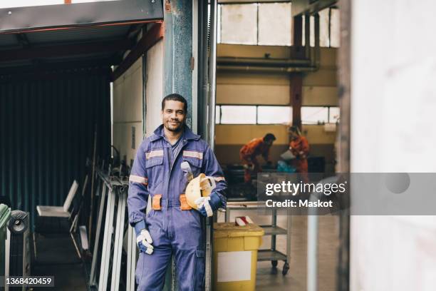 confident male worker holding hardhat in warehouse doorway - industrial labourer stock pictures, royalty-free photos & images