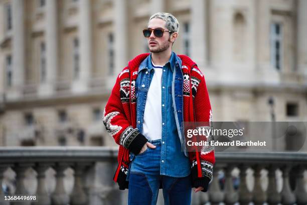 Alejandro Acero wears brown sunglasses, a white t-shirt, a blue denim jacket, a red oversized wool knit cardigan with printed patterns and stars, and...