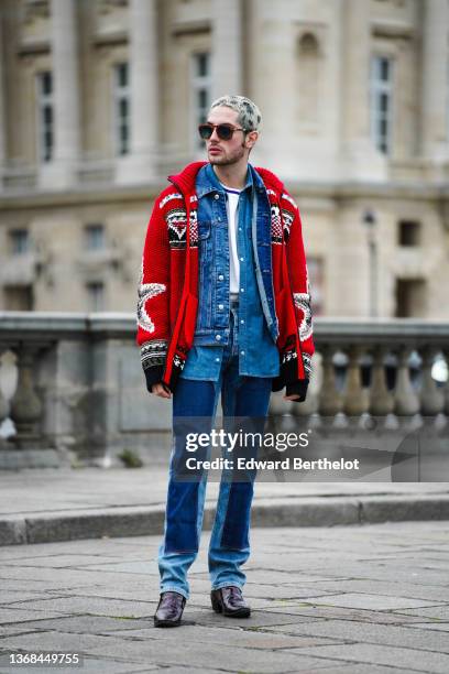 Alejandro Acero wears brown sunglasses, a white t-shirt, a blue denim jacket, a red oversized wool knit cardigan with printed patterns and stars, and...