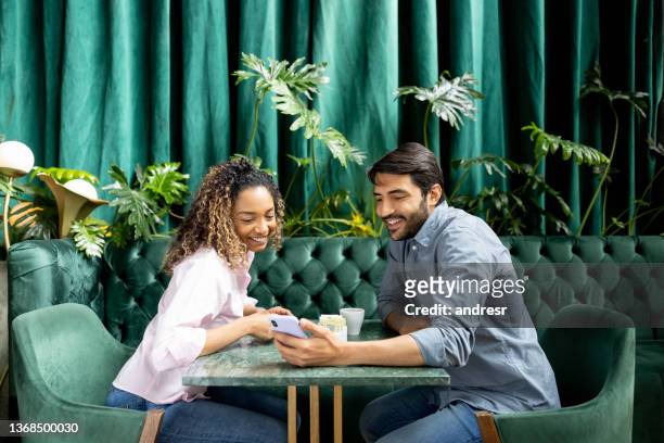 couple having coffee at a cafe and looking at social media on their cell phone - african american restaurant texting stockfoto's en -beelden
