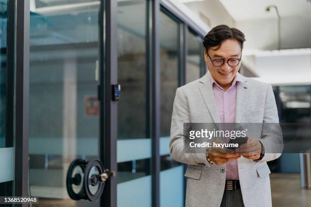 mature businessman using smartphone in office corridor - texting at work stock pictures, royalty-free photos & images