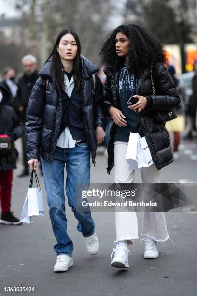 Guest wears a black t-shirt, a white striped shirt, silver earrings, a black shiny oversized puffer jacket, blue denim jeans pants, a white shopping...