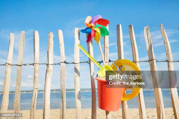 red bucket, pinwheel toy and yellow shovel hanging at a fence at the beach against blue sky - beach shovel stockfoto's en -beelden