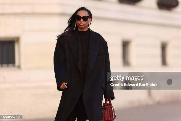 Emilie Joseph wears sunglasses, rhinestones pendant earrings, a black long knitted midi dress, a black oversized mohair winter coat from The...