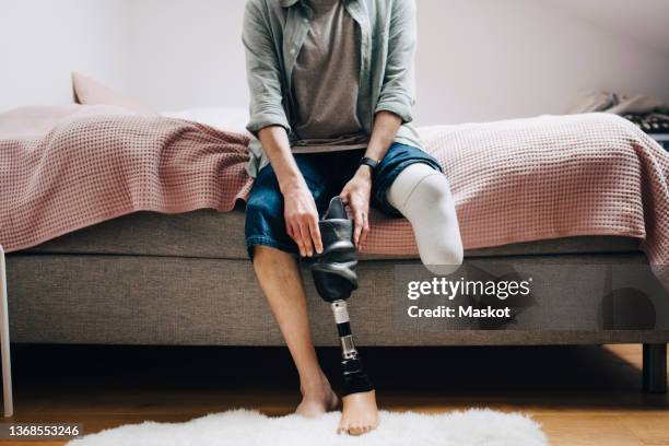 low section of disabled man with prosthetic leg sitting on bed at home - amputatie stockfoto's en -beelden