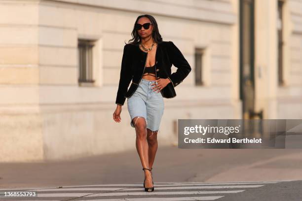 Emilie Joseph wears sunglasses, diamond earrings, a gold large chain necklace from Merbabe, a black velvet with embroidered gold borders and large...