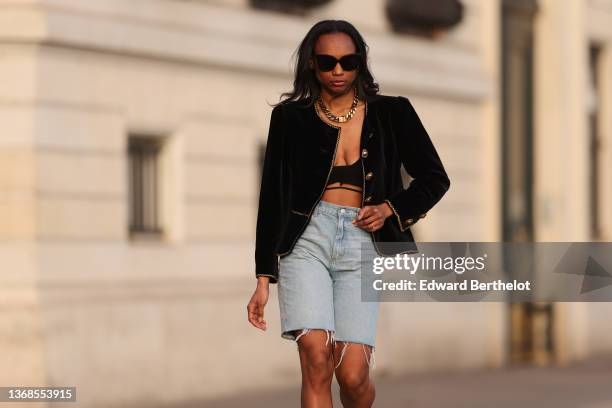 Emilie Joseph wears sunglasses, diamond earrings, a gold large chain necklace from Merbabe, a black velvet with embroidered gold borders and large...