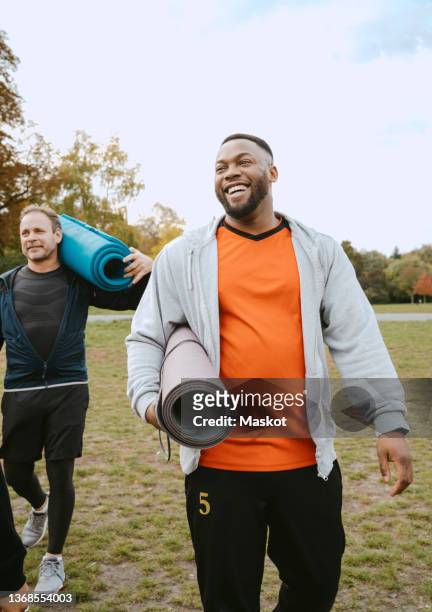 cheerful man with exercise mat walking by male friend in park - lifestyles stock pictures, royalty-free photos & images