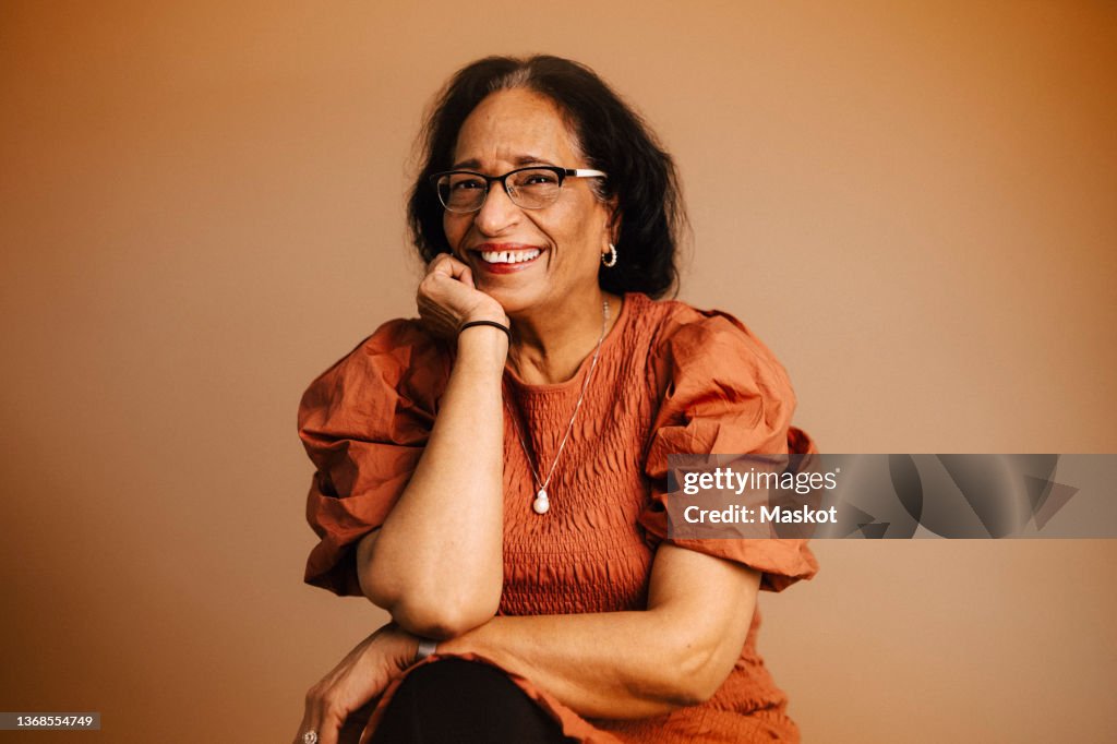 Happy senior woman with hand on chin against brown background