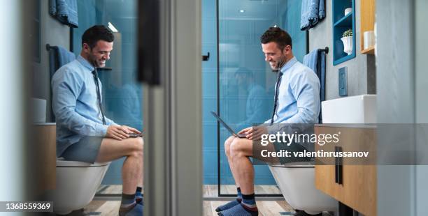 young man with shirt, sitting on the toilet, holding laptop on his knees and having online conversation in a difficult situation - trousers down stock pictures, royalty-free photos & images