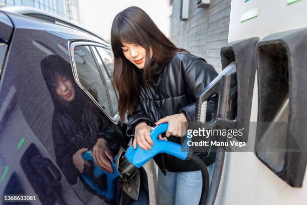 young chinese woman smiling and holding electric plug while charging her electric car - fuel efficient stock pictures, royalty-free photos & images