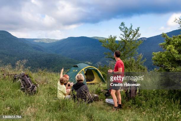 a friendly family of backpackers with children eating a snack in the camp at the tent in the evening during a long hike in the mountains. enjoy the scenic view of the mountain ranges and the beautiful cloudy sky - eco tourism stock pictures, royalty-free photos & images