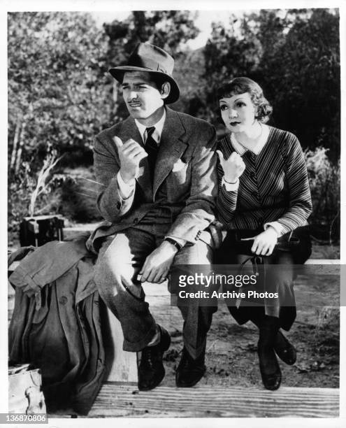 Claudette Colbert and Clark Gable sitting on a wooden fence with their thumbs pointing toward the right in a scene from the film 'It Happened One...