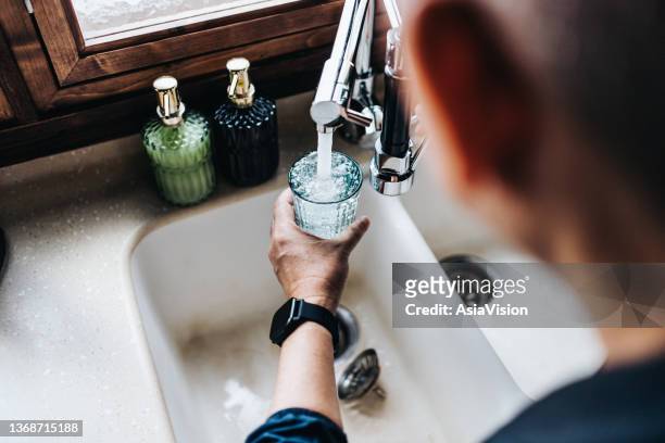 over the shoulder view of senior asian man filling a glass of filtered water right from the tap in the kitchen at home - grondstoffen stockfoto's en -beelden