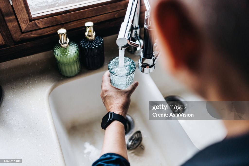 Sobre a visão do ombro do homem asiático sênior enchendo um copo de água filtrada apenas da torneira na cozinha em casa