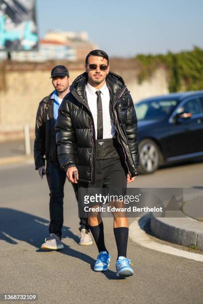 Guest wears black sunglasses, a white shirt, a black tie, a back shiny leather hoodie puffer jacket from Prada, high waist shorts from Prada, black...