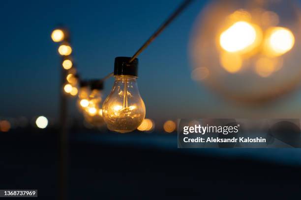 close up of the glowing light bulbs at the beach cafe terrace. sun is setting in the blurry abstract background. - bombillas fotografías e imágenes de stock