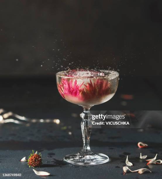 cocktail in champagne glass with flowers ice cubes and splash on dark table with petals at black wall background - cocktail stock-fotos und bilder