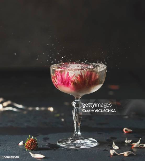 cocktail in champagne glass with flowers ice cubes and splash on dark table with petals at black wall background - cocktail stock pictures, royalty-free photos & images