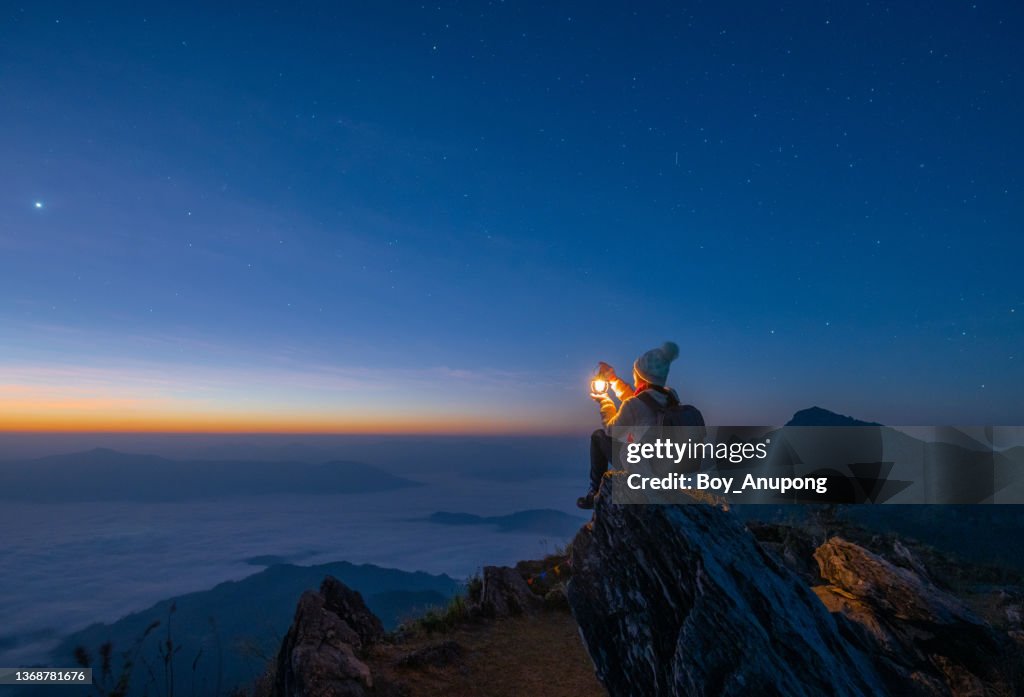 Tourist woman holding a lantern while sitting on the rock on top of Doi Pha Tang mountain a high cliff over the Thai-Laotian border located in Wiang Kaen district of Chiang Rai province of Thailand. Beautiful view before the sunrise.