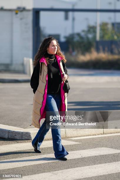 Guest wears a gold chain necklace from Dior, a black turtleneck pullover, a beige with neon pink interior long puffer jacket, blue denim flared jeans...