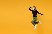 Full length of excited young arabian muslim woman wearing hijab black green clothes jumping looking far away distance isolated on yellow background studio portrait. People religious lifestyle concept.