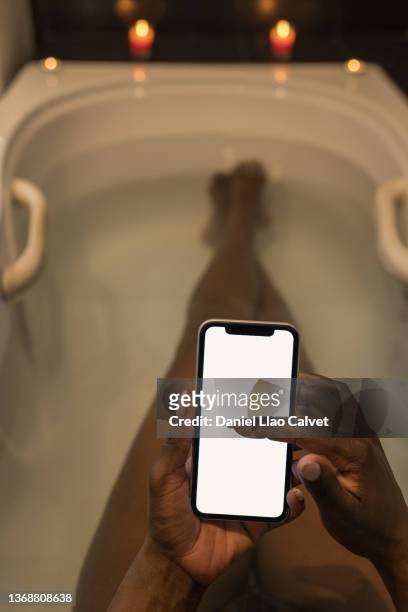 an unrecognizable african-american woman takes a hot bath while consulting her mobile phone. blank screen - african american woman bath stock pictures, royalty-free photos & images