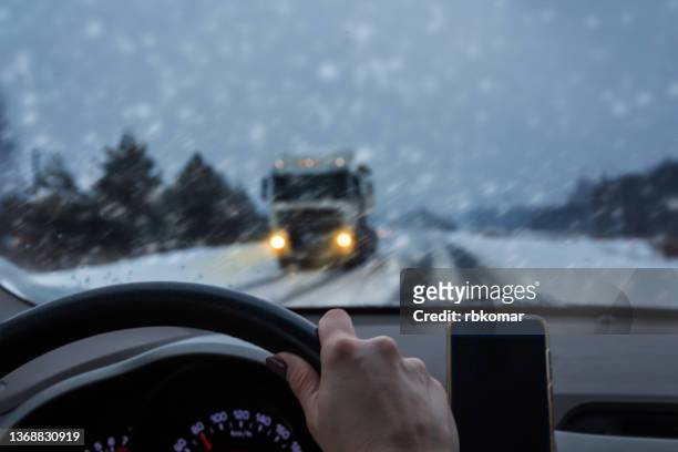 woman driver on a snowy slippery road at dusk. snowfall and blizzard while driving a car steering wheel, inside view. truck with bright headlights moving towards - winter car window stock-fotos und bilder