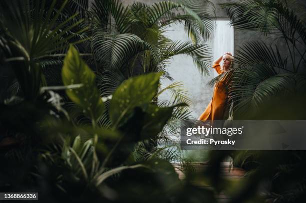 mujer apoyada en una pared de hormigón gris rodeada de verdes hojas de palma tropical, junto a la piscina. - orange dress fotografías e imágenes de stock