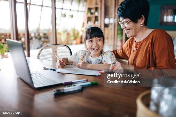 asian grandmother homeschooling her granddaughter and assist her with school work at home. little girl with headphones is studying from home and attending online school classes with laptop. e-learning, homeschooling concept - hong kong grandmother stock pictures, royalty-free photos & images