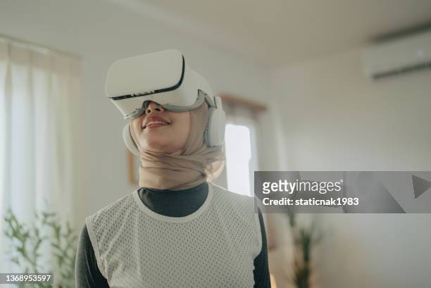 attractive islam woman wearing vr glasses sitting on sofa at home. - virtual reality simulator stock pictures, royalty-free photos & images
