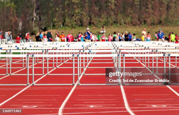 athletics hurdles placed on the tartan of the outdoor athletics track ready to start the race. in the background a large number of athletes waiting to participate - carreras de obstáculos prueba en pista fotografías e imágenes de stock