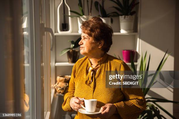ältere frau, die aus dem fenster schaut, während sie eine tasse kaffee hält - banking document stock-fotos und bilder